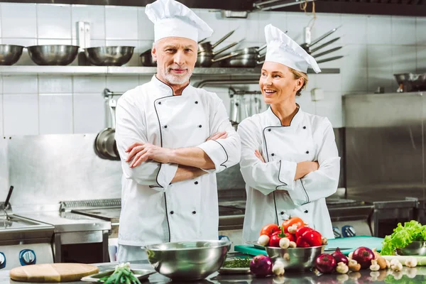 Chefs féminins et masculins en uniforme avec les bras croisés pendant la cuisson dans la cuisine du restaurant — Photo de stock