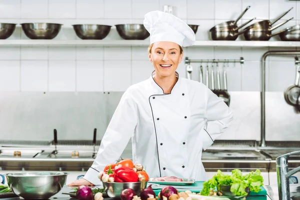 Schöne glückliche Köchin in Uniform, die beim Kochen in der Restaurantküche in die Kamera schaut — Stockfoto