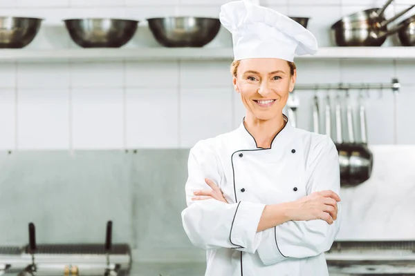 Красивий усміхнений жіночий шеф-кухар в уніформі і хет-мейт обіймає камеру на кухні ресторану — стокове фото
