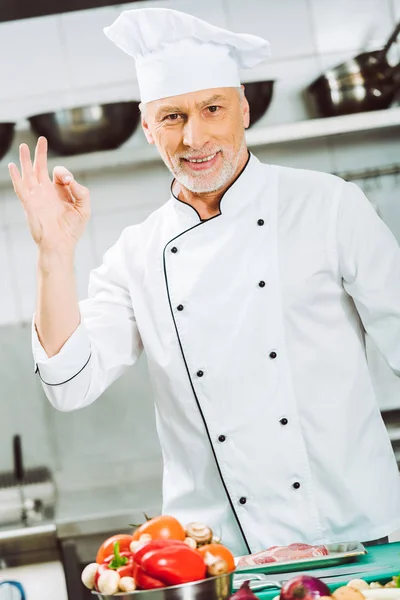 Красивый улыбающийся шеф-повар в форме смотрит в камеру и показывает знак ОК на кухне ресторана — стоковое фото
