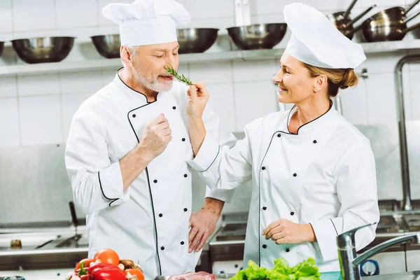 Sorridente chef donna che tiene rosmarino erba vicino all'uomo durante la cottura in cucina ristorante — Foto stock