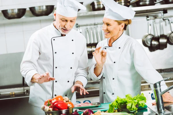 Chefs femininos e masculinos de uniforme e chapéus conversando enquanto cozinham na cozinha do restaurante — Fotografia de Stock