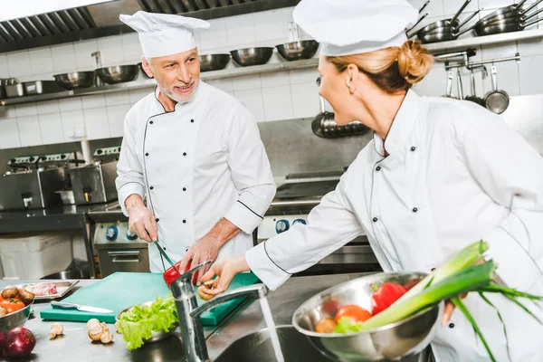Chefs femeninos y masculinos en uniforme y sombreros mirándose mientras cocinan en la cocina del restaurante - foto de stock