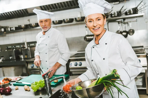 Female and male chefs in double-breasted jackets and hats looking at camera while cooking in restaurant kitchen — Stock Photo