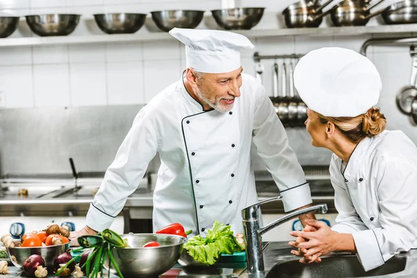 Chefs femininos e masculinos de uniforme olhando uns para os outros enquanto cozinham na cozinha do restaurante — Fotografia de Stock