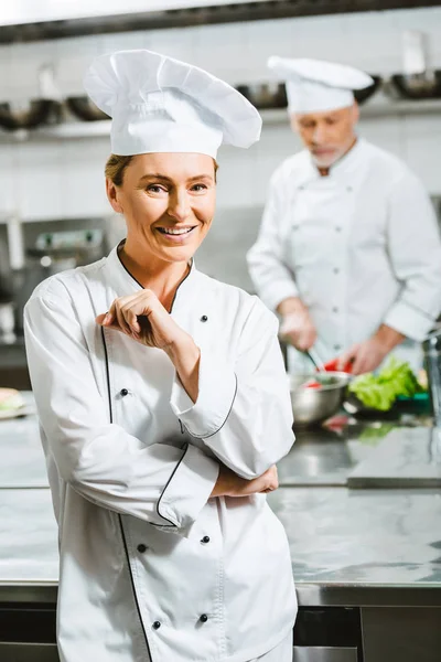 Schöne lächelnde Köchin in Uniform und Hut, die in der Restaurantküche in die Kamera schaut — Stockfoto