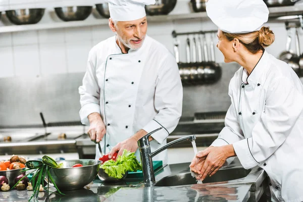 Foyer sélectif des femmes et des hommes chefs en uniforme regardant les uns les autres tout en cuisinant dans la cuisine du restaurant — Photo de stock