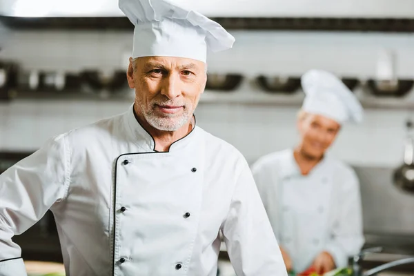 Schöner männlicher Koch in Uniform und Mütze, der in der Restaurantküche mit Kopierraum in die Kamera schaut — Stockfoto