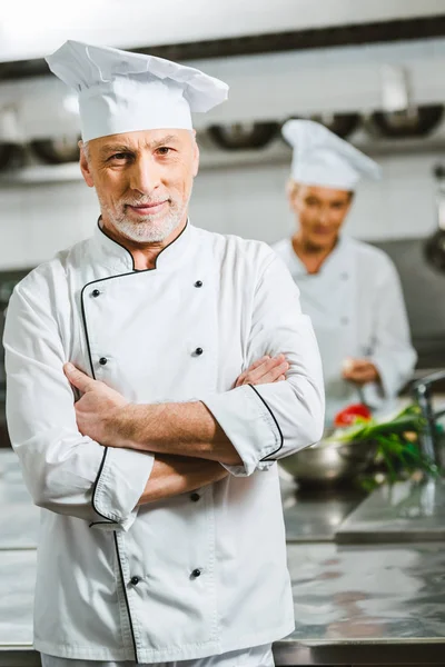 Schöner männlicher Koch in Uniform mit verschränkten Armen, der in der Restaurantküche in die Kamera blickt — Stockfoto