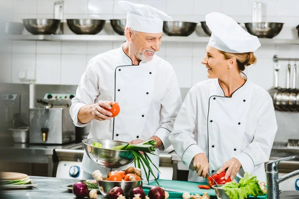 Sorrir chefs femininos e masculinos de uniforme olhando uns para os outros enquanto cozinham na cozinha do restaurante — Fotografia de Stock