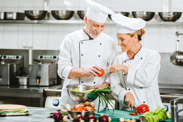 Chef sorridenti di sesso femminile e maschile in giacche e cappelli doppiopetto che cucinano nella cucina del ristorante — Foto stock