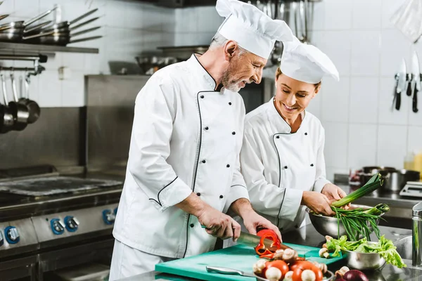 Smiling female and male chefs in uniform and hats cooking in restaurant kitchen — Stock Photo