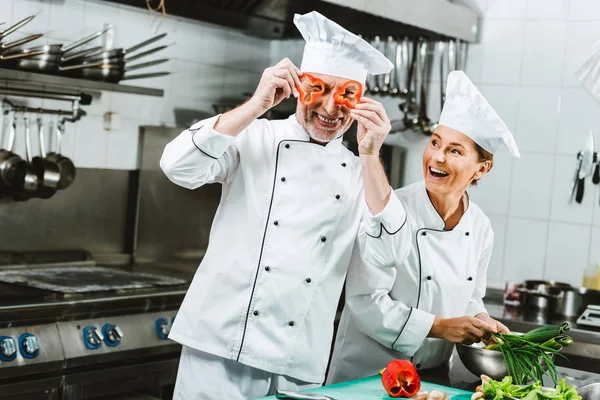 Усміхнена жінка готує, дивлячись на шеф-кухаря, який тримає шматочки перцю перед обличчям на кухні ресторану — стокове фото