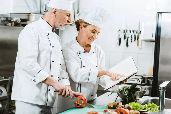 Selective focus of female and male chefs in uniform using recipe book during cooking in restaurant kitchen — Stock Photo