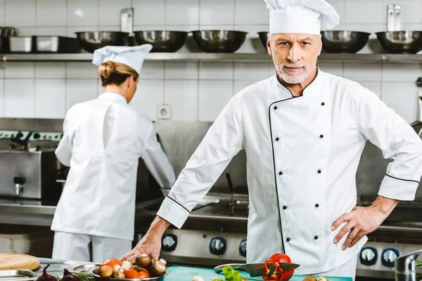 Cher chef masculin en uniforme regardant la caméra pendant la cuisson dans la cuisine du restaurant avec une collègue féminine sur fond — Photo de stock