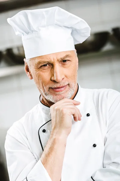 Selektiver Fokus des gut aussehenden männlichen Chefkochs, der sein Kinn mit der Hand stützt und in der Restaurantküche in die Kamera blickt — Stockfoto