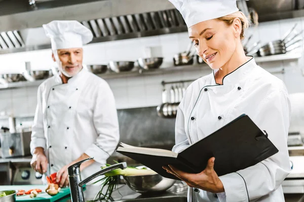 Female chef in uniform reading recipe book while colleague cooking on background in restaurant kitchen — Stock Photo