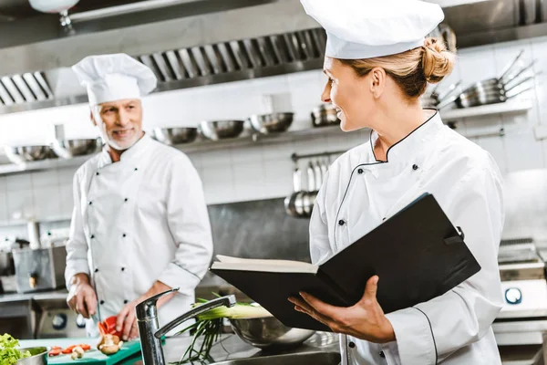 Mujer chef en uniforme celebración de libro de recetas mientras colega de cocina en el fondo en la cocina del restaurante - foto de stock