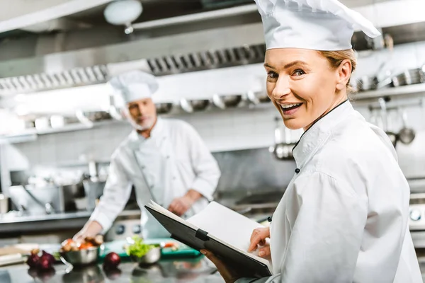 Hermosa mujer chef en uniforme celebración de libro de recetas y mirando a la cámara en la cocina del restaurante con colega en el fondo - foto de stock