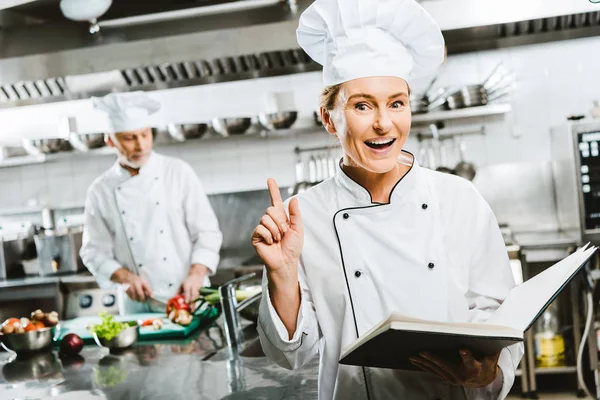 Красивая женщина шеф-повар в униформе держит книгу рецептов и делает жест идеи в кухне ресторана с коллегой на заднем плане — стоковое фото