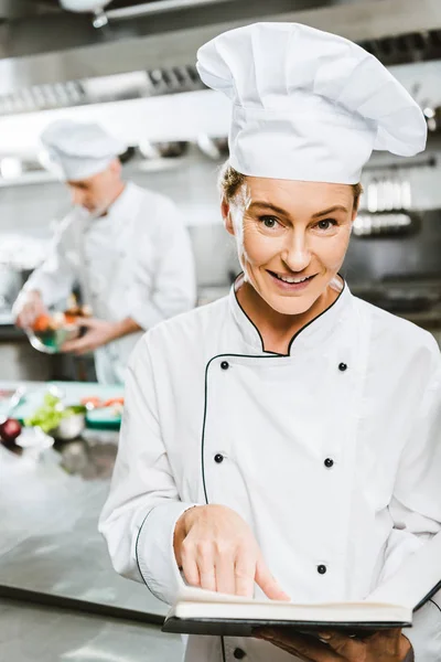 Красивая женщина шеф-повар в униформе проведение книгу рецептов и смотреть на камеру в кухне ресторана — стоковое фото