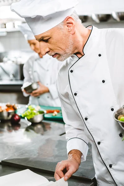 Chef masculino en uniforme y sombrero libro de recetas de lectura en la cocina del restaurante - foto de stock