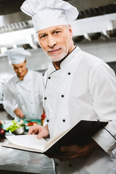Chef masculino en uniforme y sombrero mirando a la cámara y sosteniendo libro de recetas en la cocina del restaurante - foto de stock