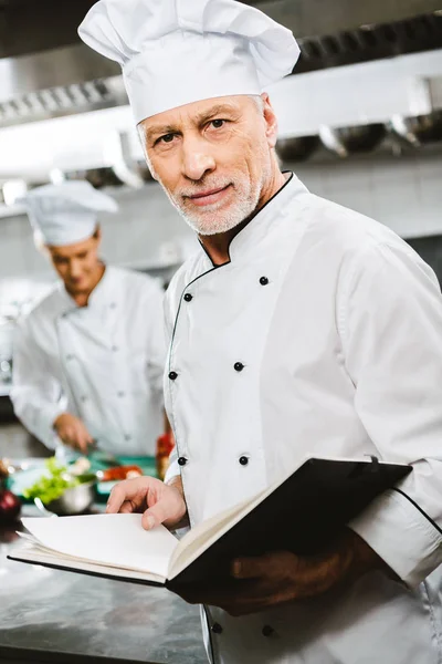 Chef masculino en uniforme y sombrero mirando a la cámara y sosteniendo libro de recetas en la cocina del restaurante — Stock Photo