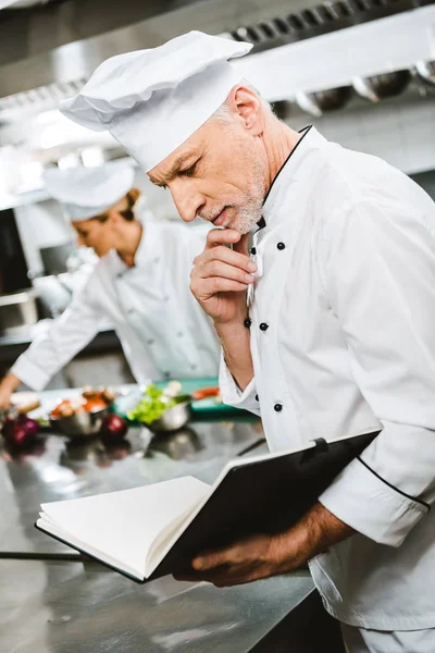 Handsome thoughtful male chef propping chin with hand while reading recipe book in restaurant kitchen — Stock Photo