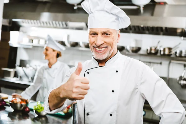 Enfoque selectivo de guapo y sonriente chef masculino en uniforme mirando a la cámara y mostrando el pulgar hacia arriba en la cocina del restaurante - foto de stock