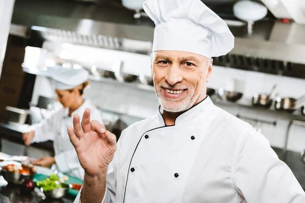 Enfoque selectivo de guapo y sonriente chef masculino en uniforme mirando a la cámara y mostrando signo de ok en la cocina del restaurante — Stock Photo