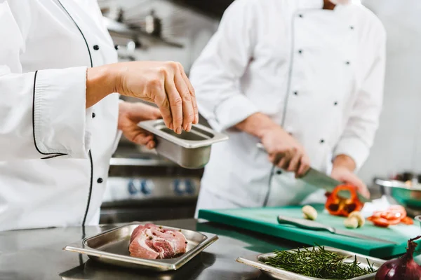 Cropped view of female chef seasoning meat while man cooking on background in restaurant kitchen — Stock Photo