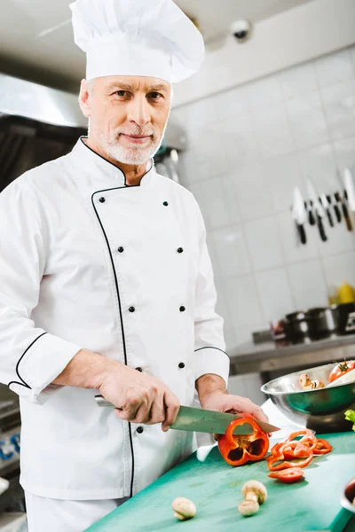 Männlicher Koch in Uniform blickt in die Kamera, während er in der Restaurantküche Pfeffer schneidet — Stockfoto