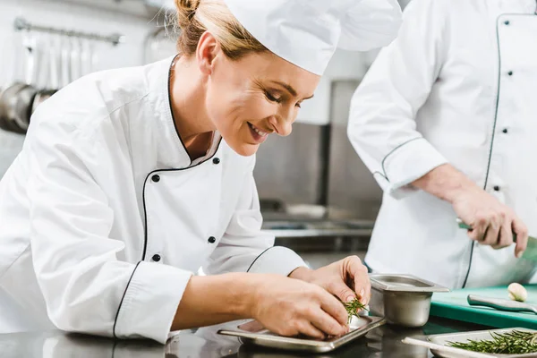 Beautiful smiling female chef in uniform decorating dish with herb in restaurant kitchen — Stock Photo