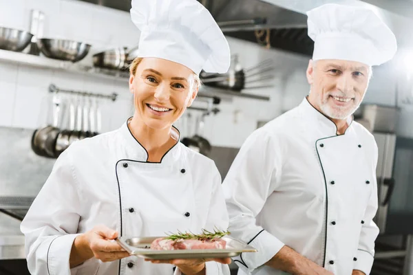 Female and male chefs in uniform with meat steak on plate looking at camera in restaurant kitchen — Stock Photo