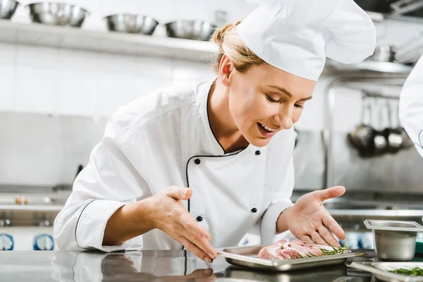 Hermosa hembra sonriente chef en uniforme sosteniendo plato con plato de carne en la cocina del restaurante - foto de stock