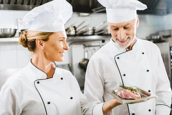 Chef sorridente in uniforme che presenta il piatto di carne al collega nella cucina del ristorante — Foto stock