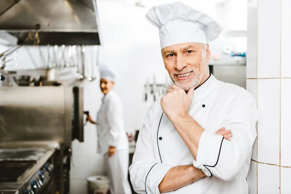 Sonriente macho guapo chef apoyando la barbilla con la mano y mirando a la cámara en la cocina del restaurante - foto de stock