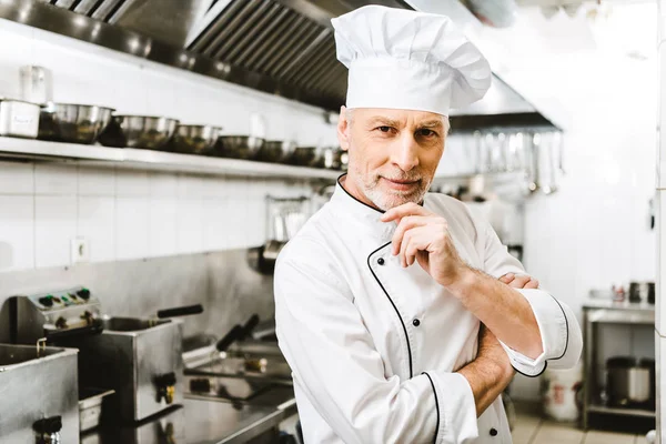 Guapo macho chef en uniforme y gorra tocando barbilla andf mirando cámara en restaurante cocina - foto de stock