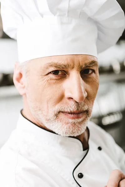 Retrato de guapo chef masculino en uniforme y gorra mirando a la cámara en la cocina del restaurante - foto de stock