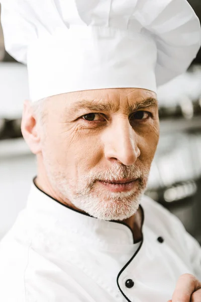 Portrait of handsome male chef in uniform and cap looking at camera in restaurant kitchen — Stock Photo