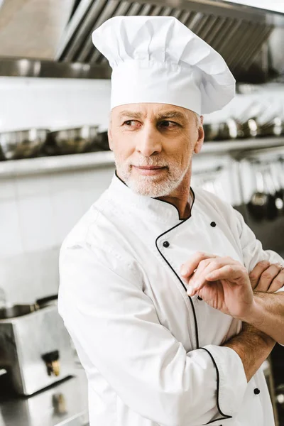 Guapo chef masculino en uniforme y gorra mirando hacia otro lado en la cocina del restaurante - foto de stock