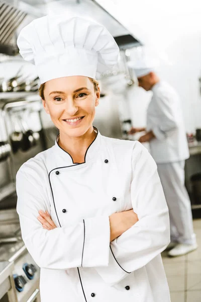 Beautiful smiling female chef in uniform with arms crossed looking at camera in restaurant kitchen — Stock Photo