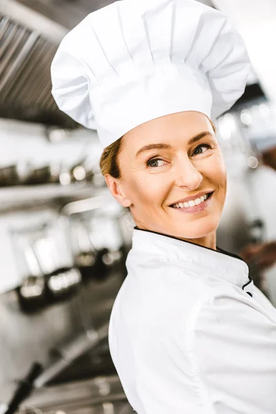 Селективный фокус красивой улыбающейся женщины-шеф-повара в кепке, смотрящей в сторону на кухне ресторана — стоковое фото
