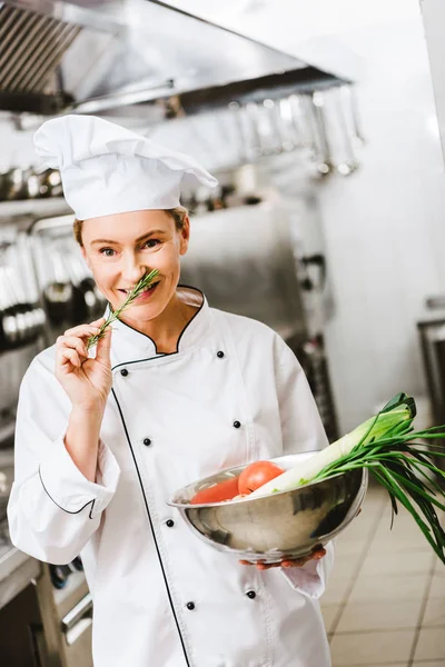 Красивая улыбающаяся женщина-шеф-повар в форме держит розмарин и миску с овощами на кухне ресторана — стоковое фото