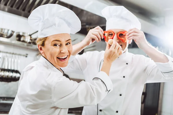 Cuoca che guarda la macchina fotografica mentre chef maschio tiene le fette di pepe davanti al viso nella cucina del ristorante — Foto stock