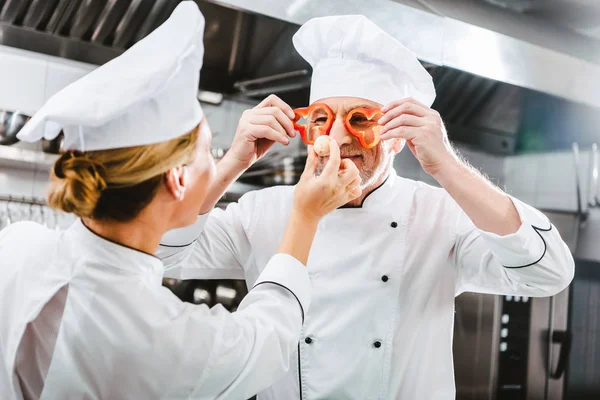 Жінка готує, дивлячись на чоловічий шеф-кухар, що тримає шматочки перцю перед обличчям на кухні ресторану — стокове фото