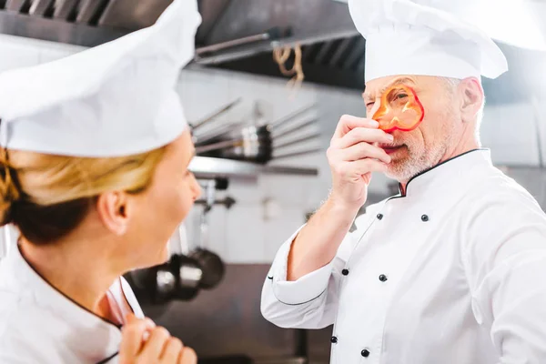 Жінка готує, дивлячись на чоловічий шеф-кухар, який тримає шматочок перцю перед обличчям на кухні ресторану — стокове фото