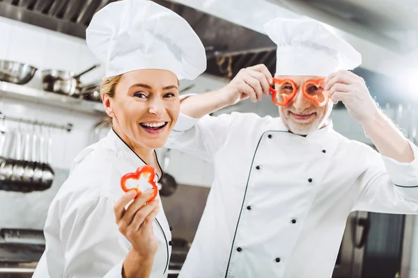 Cocinera femenina feliz mirando a la cámara mientras chef masculino sosteniendo rodajas de pimienta en frente de la cara en la cocina del restaurante - foto de stock