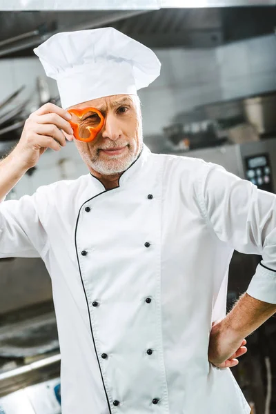 Chef maschio in uniforme e cappello con fetta di pepe davanti al viso nella cucina del ristorante — Foto stock